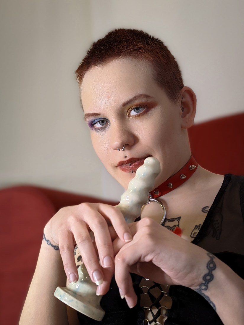 Pinja katsoo kasvoista otetussa puolilähikuvassa hiukan ohi kamerasta käsissään valkoinen keraaminen dildo.