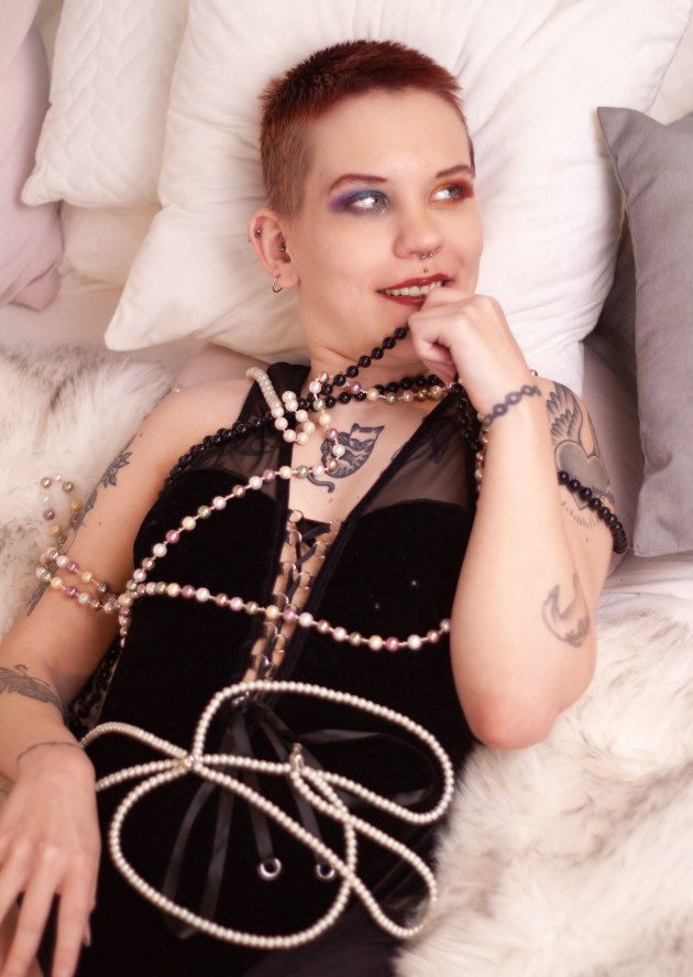 Pinja makaa sängyllä mustassa bodyssa ja leikkii kädessä olevalla helminauhalla.