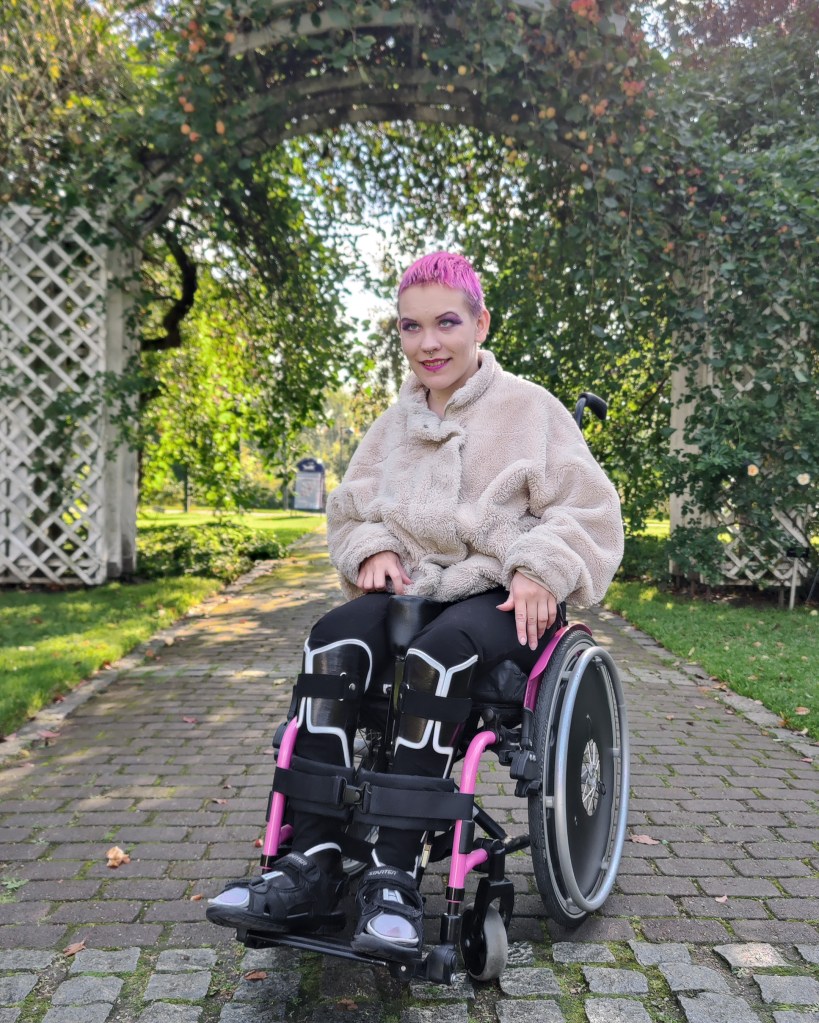 Pinja istuu pyörätuolissa melkein syksyisessä puutarhassa katsoen kameraan, yllään valkoinen pörrötakkia.