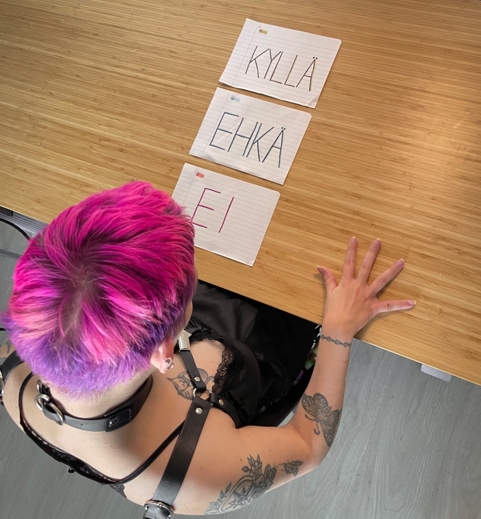 Ylhäältä päin otettu kuva, jossa istun pöydän ääressä ja edessäni on allekkain laput, joissa lukee KYLLÄ, EHKÄ, EI. Kirkkaan väriset pinkki-violetit hiukseni näkyvt hyvin.