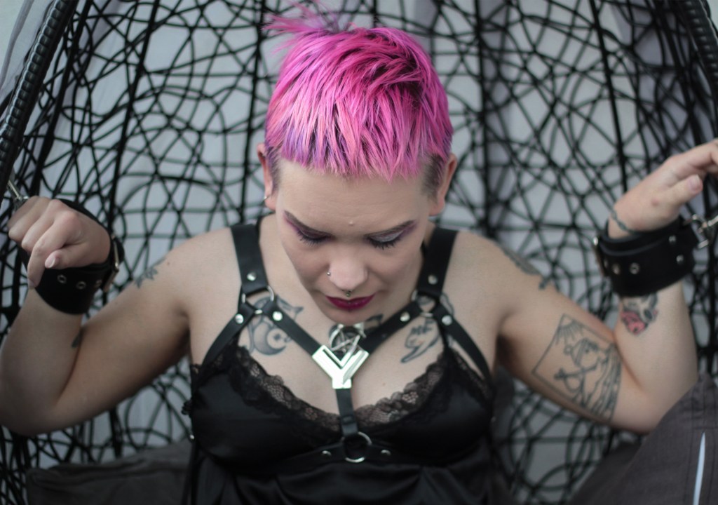 Puolikuva, jossa istun roikkuvassa korituolissa ja katson alaspäin. Takanani näkyvät tuolin mustat punokset, jotka nousevat pääni yläpuolelle. Ranteissani on nahkaiset kahleet, jotka ovat kiinni tuolin reunoissa.