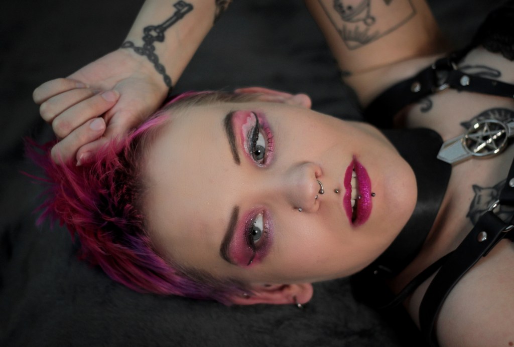 Pinja makaa selällään lähikuvassa, jonka keskiössä ovat hänen kasvonsa. Tukka on kirkkaan vaaleanpunainen, samoin silmämeikki ja huulet. Pinjalla on päällään nahkainen harnes ja hän katsoo kameran ohi.