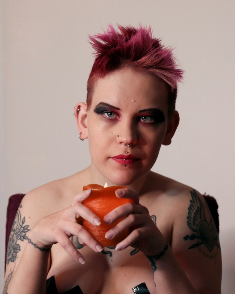 Kuvassa PInja katsoo kameraan ylävartalo paljaana. Hänellä on voimakas punamusta silmämeikki ja käsissään hän pitelee punaista kynttilää. Nännit on peitetty mustalla teipillä, jonka yläreunat on näkyvissä.