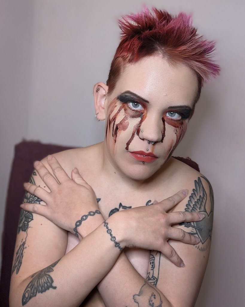 Voimakkaasti valaistussa kuvassa Pinja on peittänyt paljaan ylävartalonsa käsillään. Meikki on sotkuinen, ja kasvoilla on tekoverta, joka näyttää siltä kuin se valuisi silmistä.