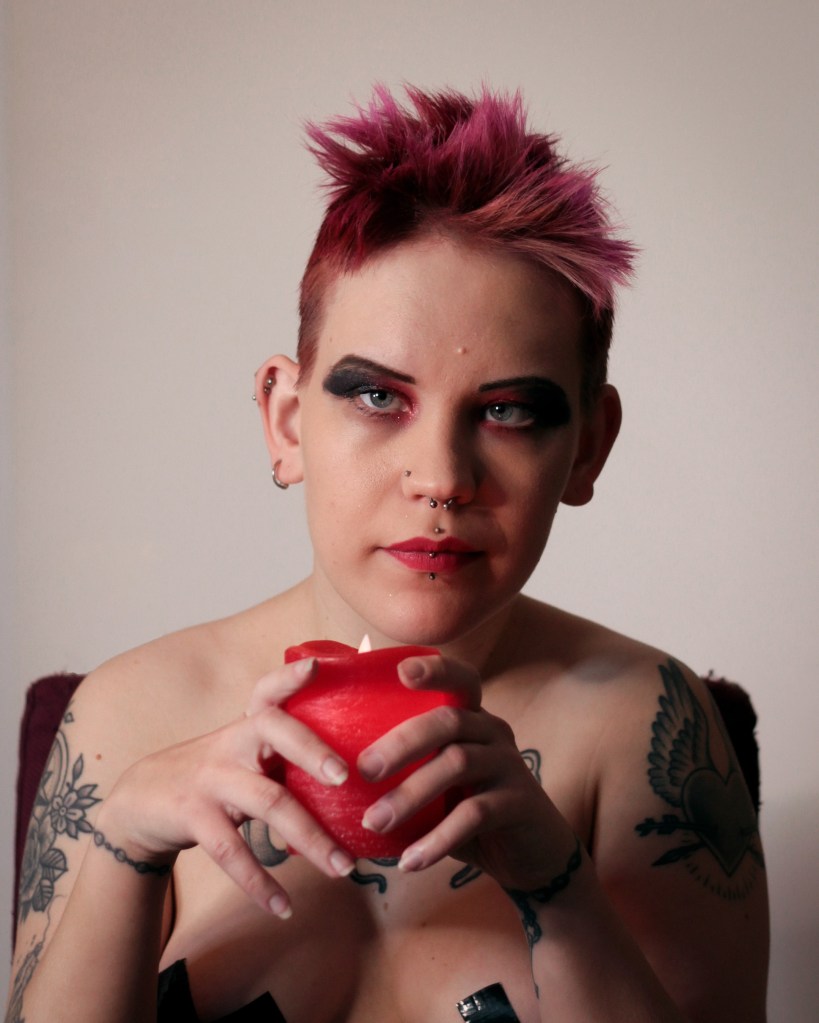 Pinja istuu tuolilla ilman paitaa ja katsoo kameraan kädessään punainen kynttilä.