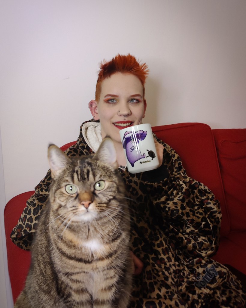 Pinja istuu punaisella sohvalla päällään jättimäinen gebardi-kuvioinen huppari ja kädessään muki jossa mökösieni potkii käpyä. Vieressä sohvan käsinojalla istuu kissa.