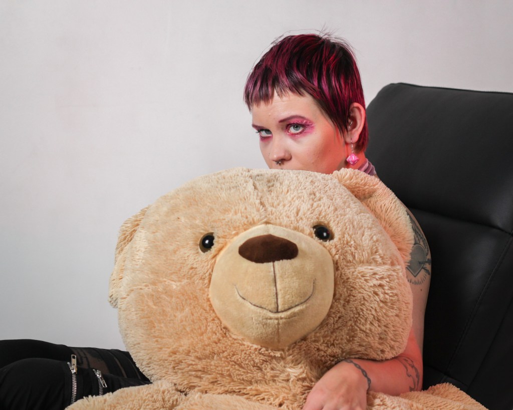 Sivukuva Pinjasta, joka istuu terapiatuolin kaltaisella virityksellä. Iso osa Pinjasta on piilossa jättikokokoisen nallen, jota hän halaa, takana. 