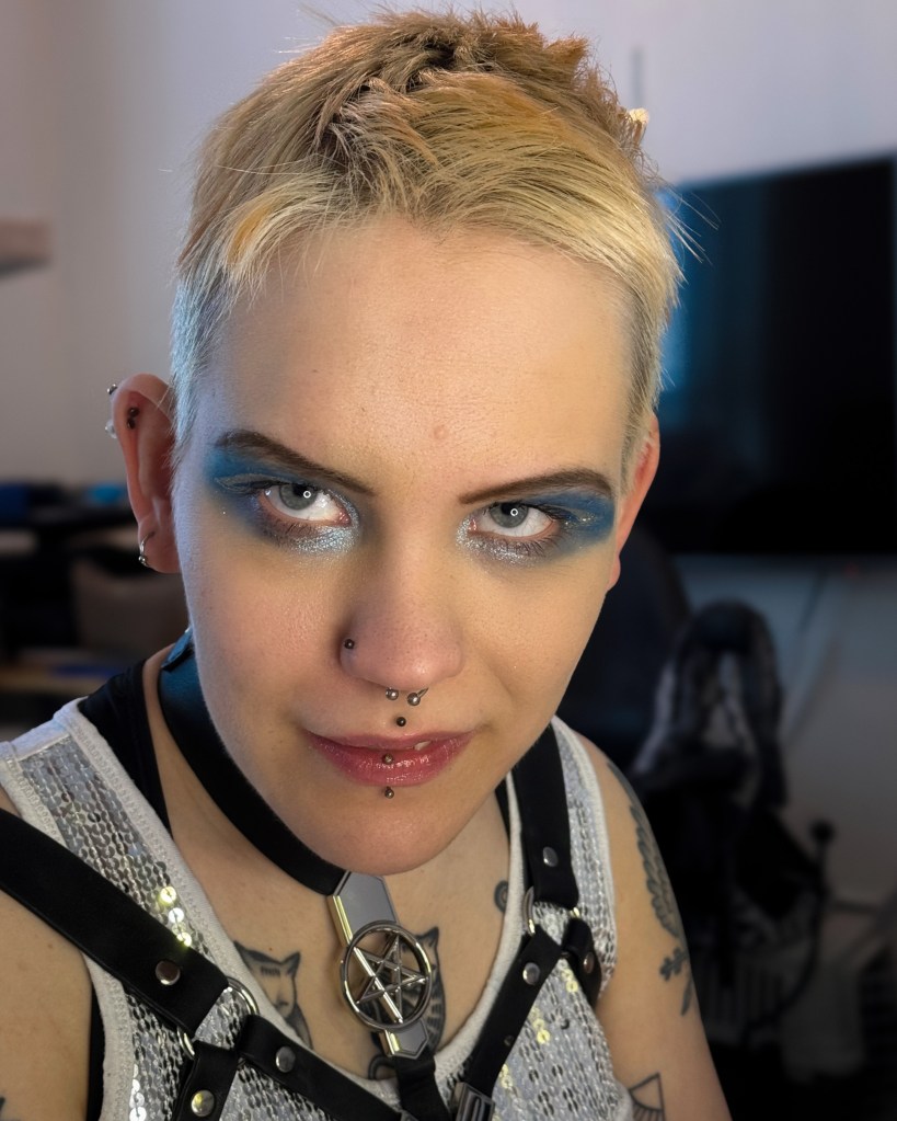 Lähikuva Pinjan kasvoista. Hänellä on vaaleat lyhyet hiukset ja sini-hopeinen silmämeikki. Katse on intensiivinen.