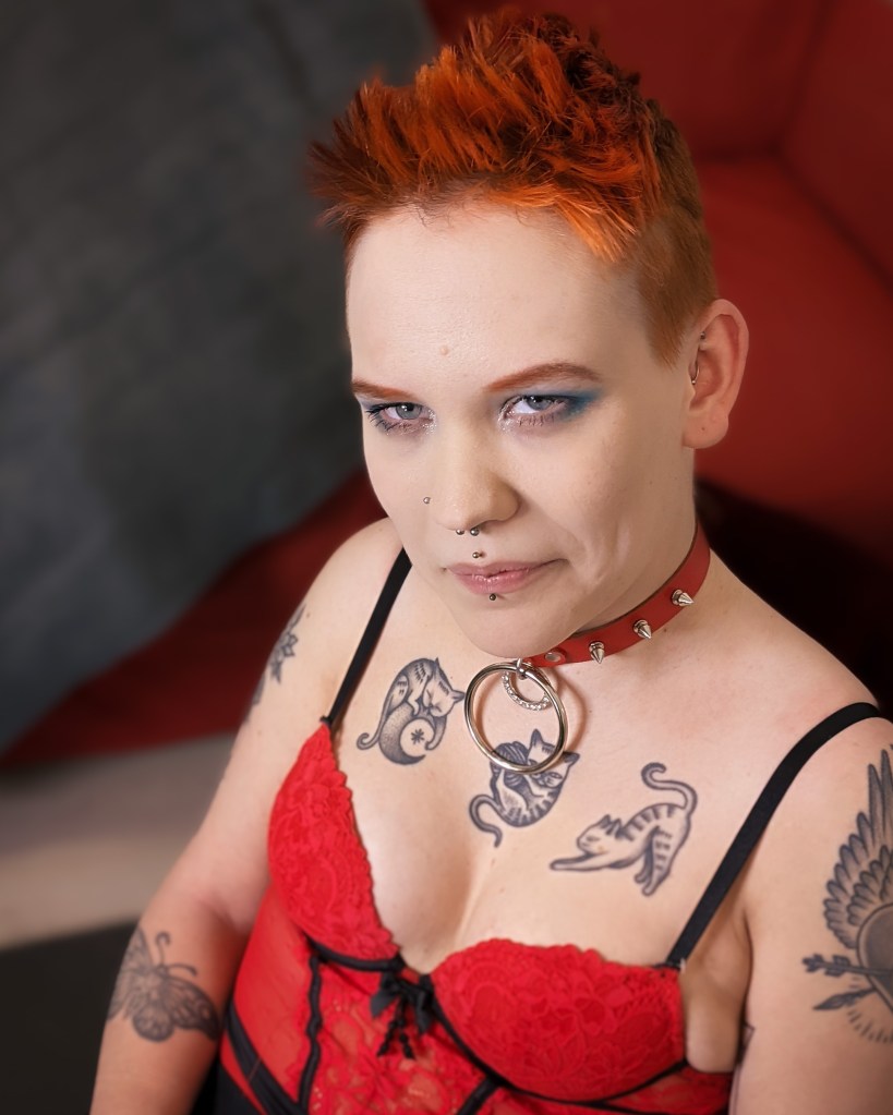 Pinja istuu ja katsoo kameran ohi. Hänellä on oranssit lyhyet hiukset ja voimakas meikki. Päällään Pinjalla on punainen alusasu ja kaulassa punainen panta jossa on piikkejä.