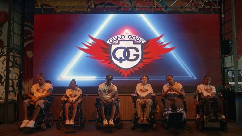 Quad Gods -joukkueen pelaajat istuvat pyörätuoleissaan, taustallaan valkokankaalle heijastettu joukkueen logo.