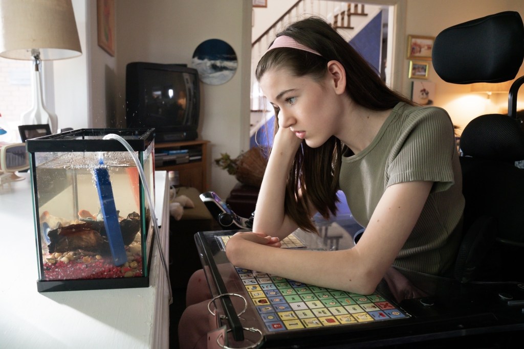 Melody istu sähköpyörätuolissaan, johon kiinnitetyllä pöydällä näkyvät kommunikaatiokortit. Hän tuijottaa tylsistyneen tai surullisen näköisenä pienessä akvaariossa uiskentelevaa lemmikkikultakalaansa Ollieta.