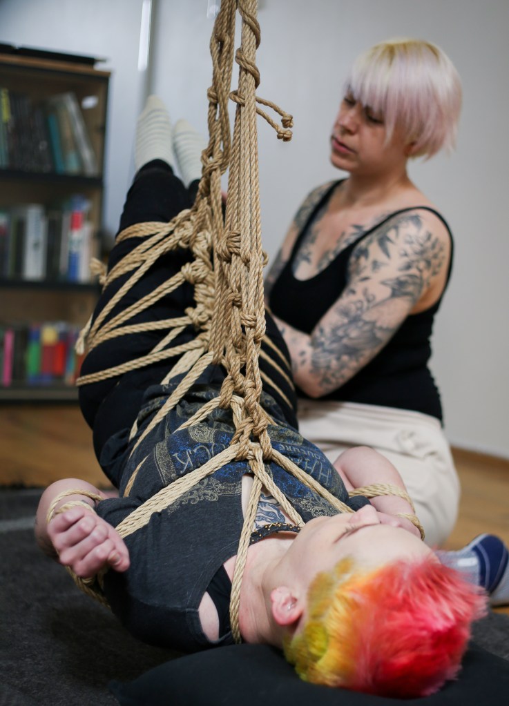 Pinja roikkuu suspensiossa. Köydet kiertävät hänen koko vartalonsa ympäri ja kiinnittyvät ylös kattoon, nostaen hänet lähes kokonaan ilmaan. Pinjan jalat ovat korkealla ilmassa ja pää lepää kevyesti lattialla olevalla tyynyllä. Pinjan takana Amy tarkastaa köysiä keskittyneen näköisenä.