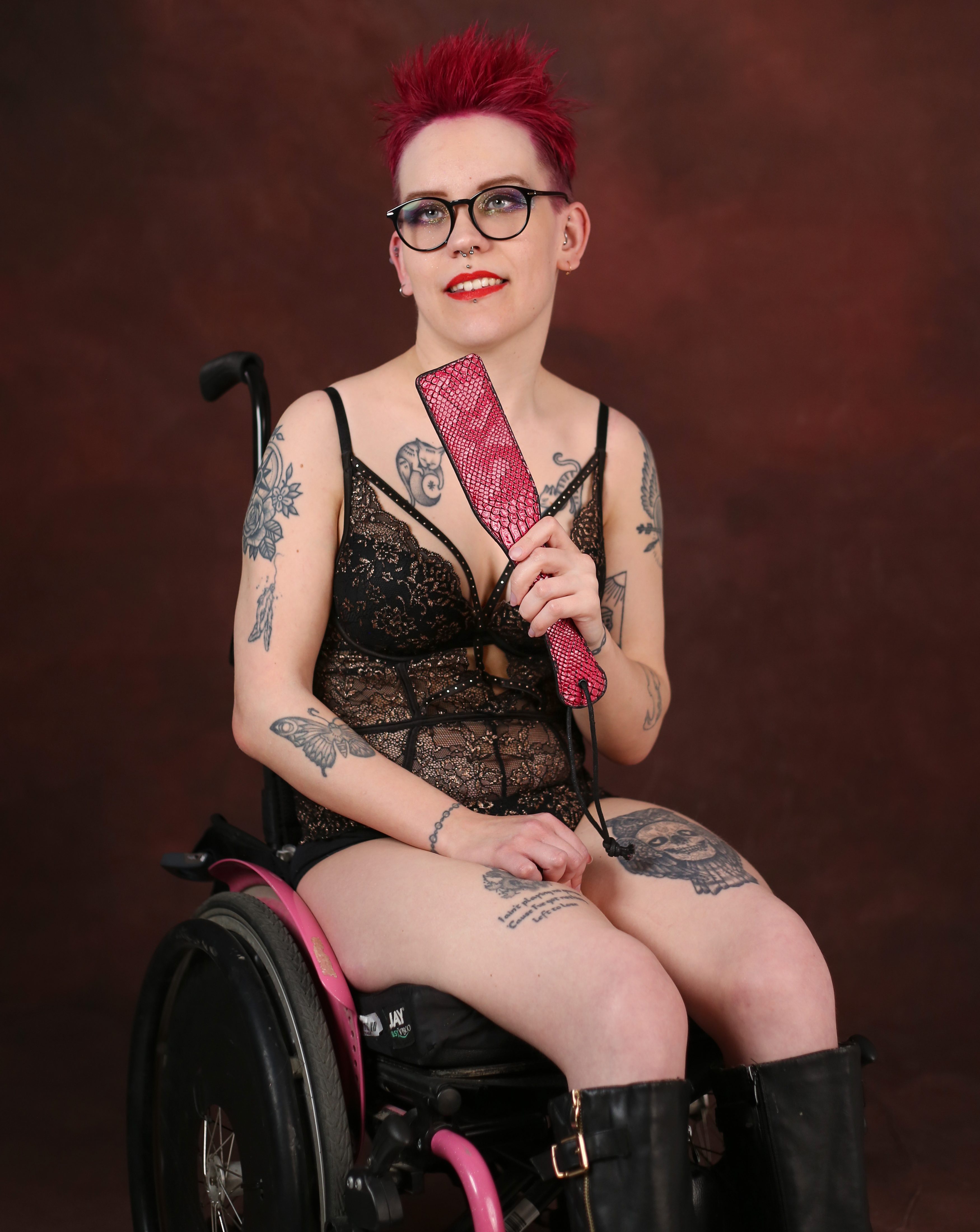Pinja, punahiuksinen tatuoitu nuori nainen, istuu pyörätuolissa pukeutuneena musta-kultaiseen bodyyn. Hän katselee hymyillen kameraan, kädessään pinkki paddle. 