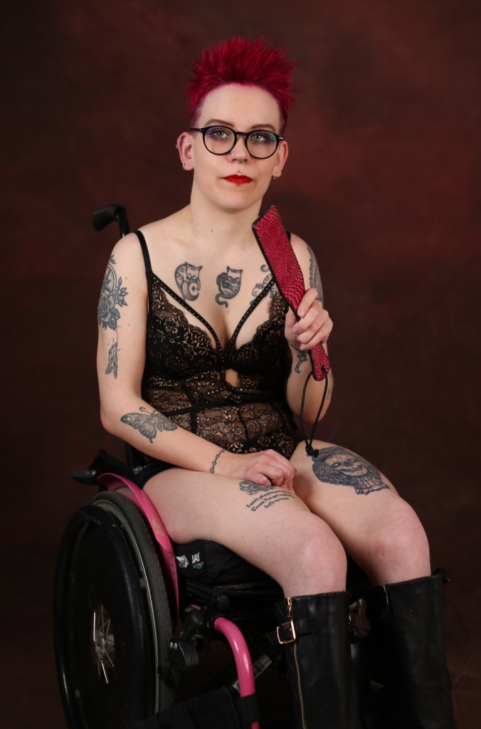 Pinja, punahiuksinen tatuoitu nuori nainen, istuu pyörätuolissa pukeutuneena musta-kultaiseen bodyyn. Hän katselee vakavana kameraan, kädessään pinkki paddle. 